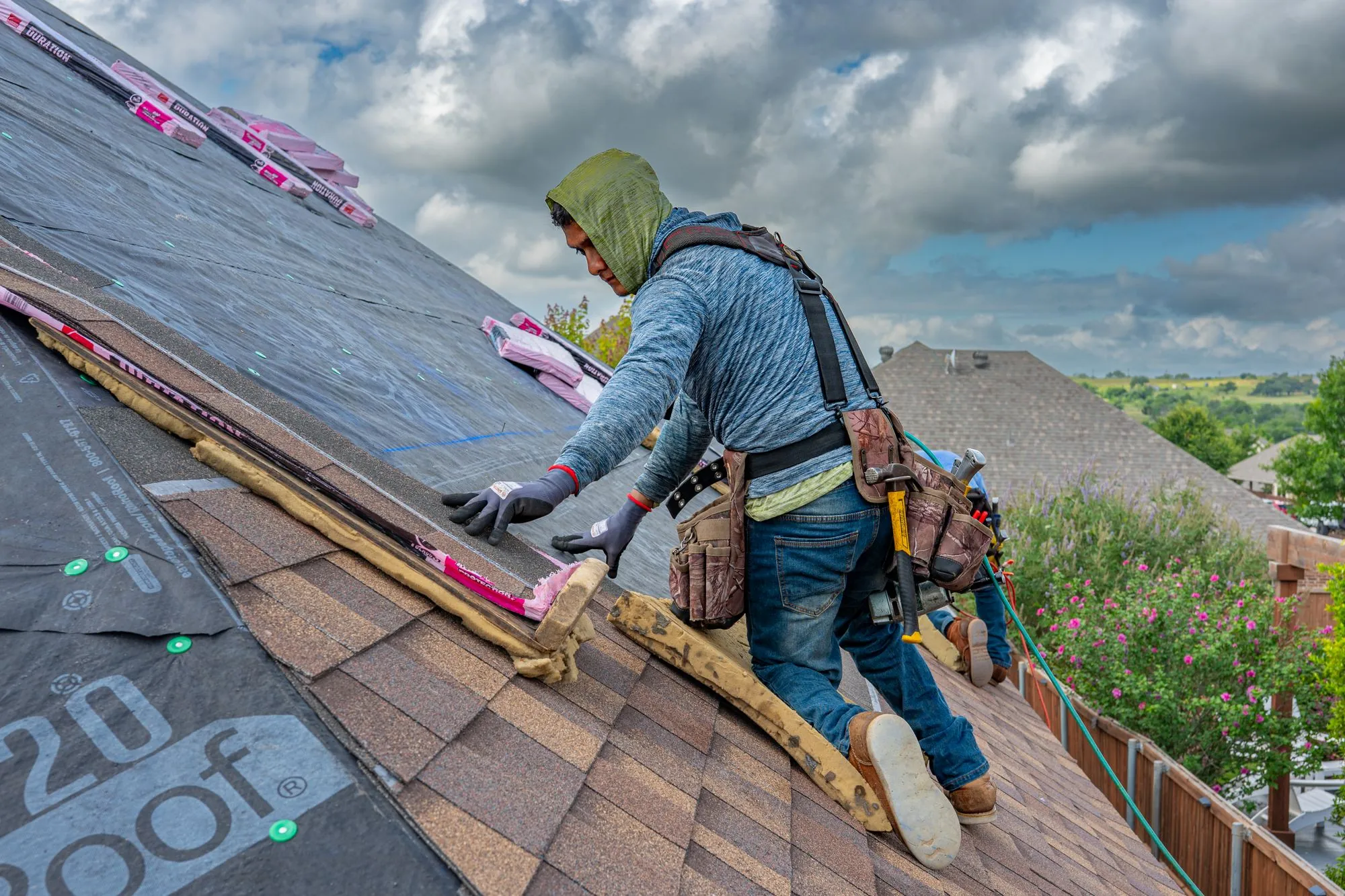 A roofer kneeling to lay a course of shingles under a cloudy sky
