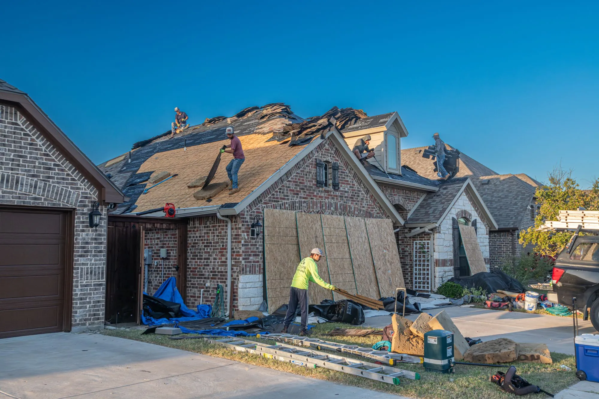 A crew re-covering the roof of a brick house, materials stacked on the drive